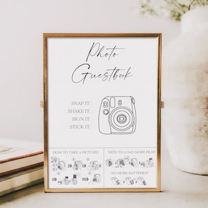 Op de afbeelding: Een wit bord met zwarte tekst en een zwart-witte illustratie van een instant camera. Het bord staat "Photo Guestbook" en bevat instructies voor het gebruik van de camera: "Snap it. Shake it. Sign it. Stick it." Het bord bevat ook instructies over hoe je een foto kunt maken en wat je moet doen als je meer film moet laden of als de batterijen leeg zijn.