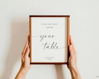 May include: A framed sign with the text "Locate your name and find your table" in elegant script. The sign is held by two hands. The bottom of the sign reads "Lily & George" and a date. The frame is brown.