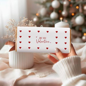 May include: A white chocolate bar with red heart designs and the text "BE MY Valentine." The bar is held in a person's hand, with a blurred background of a Christmas tree and decorations.