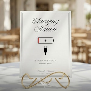 May include: A white sign with black text that reads "Charging Station" and "Recharge your devices here." The sign has a battery icon and a charging cable icon. It is displayed in a gold metal holder.