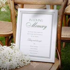 May include: A white framed sign with the words "IN LOVING Memory" and a list of names. The sign is propped on a wooden chair, with a bouquet of white flowers in front. The sign is for a wedding or memorial.