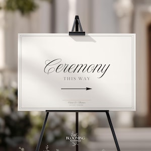 May include: A rectangular white sign with a thin black border, featuring the black text "Ceremony" in a script font, "THIS WAY" in a sans-serif font, a right-pointing arrow, and the names "Olivia & Thomas" with a date below on a black easel.