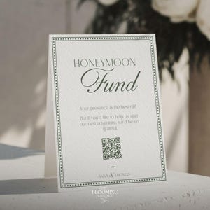 May include: A white card with the words "HONEYMOON Fund" in elegant script. The card has a decorative border and includes a QR code. Text below reads "Your presence is the best gift!" and "But if you'd like to help us start our next adventure, we'd be so grateful."