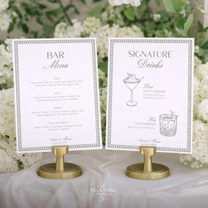 May include: Two white framed signs with black text on gold stands. One sign reads "Bar Menu" and the other "Signature Drinks". The signs are surrounded by white flowers. The bar menu lists beer, wine, spirits, and non-alcoholic drinks.