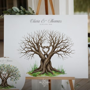 Pode incluir: Uma alternativa ao livro de visitas de casamento branco com um design de árvore em aquarela. A árvore tem troncos entrelaçados formando um coração, com os nomes "Olivia & Thomas" e a data "June 6th, 2020" impressos no topo. Um livro de visitas redondo menor também é visível.