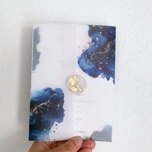 Peut inclure: Une invitation avec un motif aquarelle dans les tons bleu et or. L'invitation est scellée avec un cachet de cire avec des accents de feuilles d'or. L'invitation est tenue dans une main.