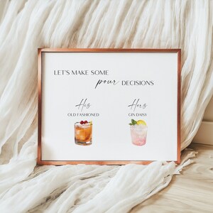 May include: A framed print with a copper-colored border. The white background features the text "LET'S MAKE SOME pour DECISIONS" and illustrations of cocktails: an Old Fashioned and a Gin Daisy. The print is displayed on a white fabric.