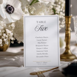 May include: A white table card with the word "TABLE" above the word "Five" in elegant script. Below are guest names. The card is set on a white tablecloth, with white flowers and a gold candlestick in the background.