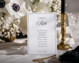 May include: A white table card with the word "TABLE" above the word "Five" in elegant script. Below are guest names. The card is set on a white tablecloth, with white flowers and a gold candlestick in the background.