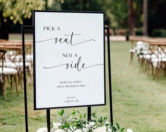 May include: A white sign with black text that reads "Pick a seat, not a side. We're all family, once the knot is tied. With love, Molly & Andy." The sign is on a black metal stand and is surrounded by white flowers.