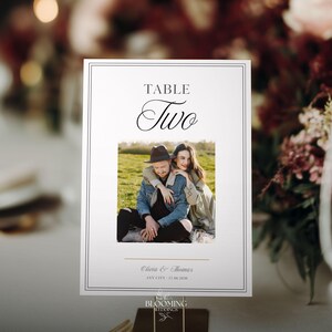 以下が含まれることがあります： 黒い縁取りとエレガントな筆記体で「TABLE Two」と書かれた白いテーブルカード。テキストの下にはカップルの写真が中央に配置されています。カードには「Olivia & Thomas」という名前と日付が記載されています。