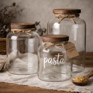 May include: Three clear glass storage jars with dark wooden lids. Each jar features a jute twine bow tied around the neck. The front-most jar has the word pasta printed in white script and a brown paper tag reading Handcrafted with love.