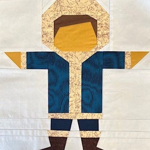 May include: A fabric quilt block featuring a stylized figure in a hooded parka. The parka is blue with gold trim, and the hood is tan. The figure has brown boots and a yellow face. The background is white.