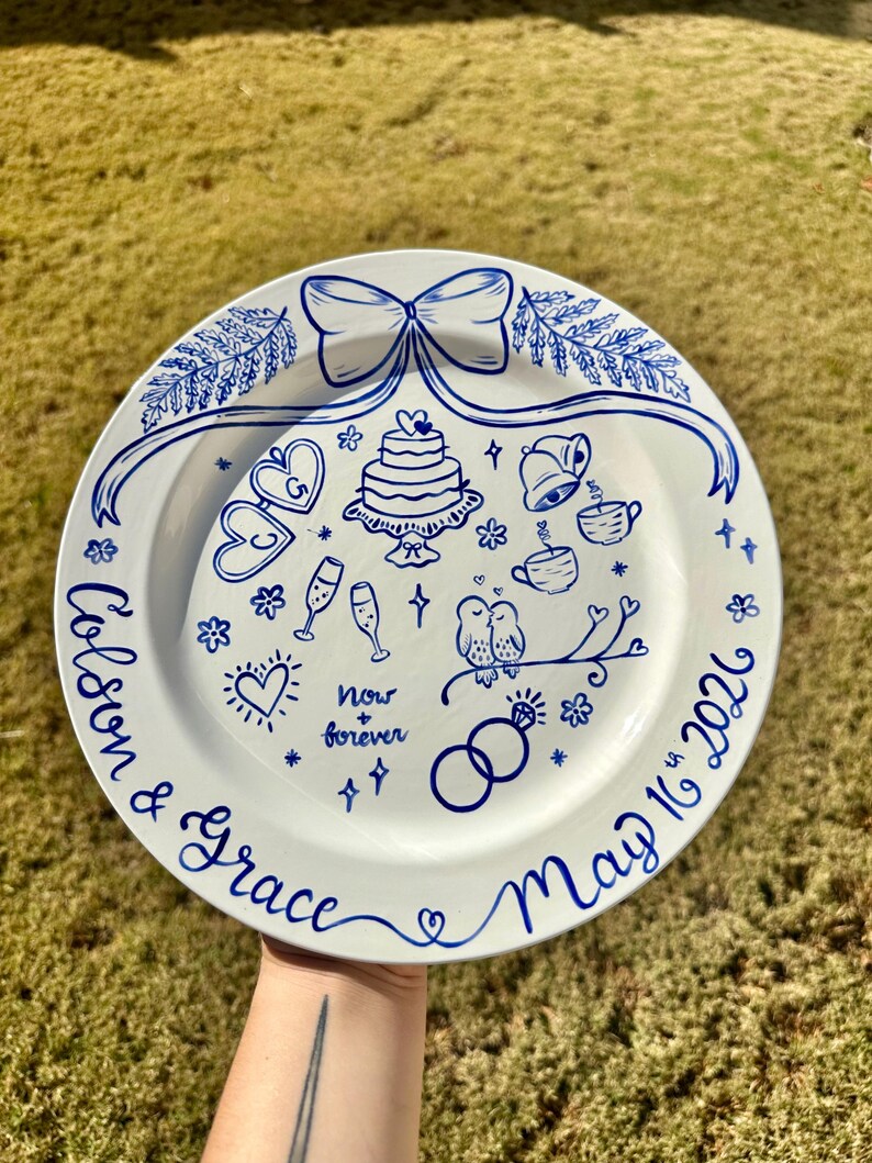以下が含まれることがあります： 手描きの青い結婚式のイラストが描かれた白いセラミックプレート。ケーキ、ハート、シャンパングラス、テキスト「Colson & Grace May 16th 2026」と「Now + Forever」が含まれています。