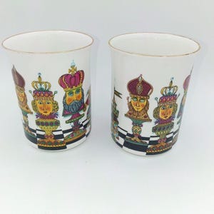 May include: Two white ceramic mugs featuring a colorful, detailed illustration of chess pieces designed as royal figures with crowns. The base of each mug displays a black and white checkered pattern, and the rims are accented with a thin gold line.