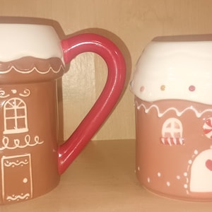 Op de afbeelding: Twee keramische mokken in de vorm van peperkoekhuisjes. Eén is bruin met witte glazuurdetails en een rode oor. De andere is roze met witte glazuurdetails, een rode oor en een zuurstokontwerp.