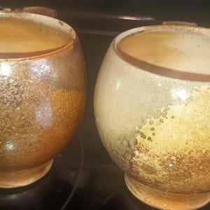 May include: Two handcrafted ceramic bowls with a speckled, textured surface. The bowls are round with a wide opening and a small, rounded base. The colors are a mix of brown, tan, and gold, with a rustic, earthy aesthetic.