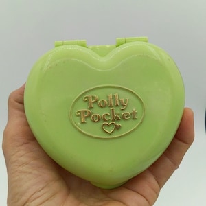 May include: A light green heart-shaped Polly Pocket compact case with a gold-colored oval emblem on the front. The emblem features the text Polly Pocket and a small heart icon. The plastic case has a smooth finish and a hinged top closure.