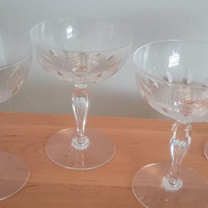 Könnte beinhalten: Vier klare Champagner-Coupe-Gläser mit geätzten floralen Designs. Die Gläser haben einen runden Fuß, einen dekorativen Stiel und eine breite, flache Schale. Die Gläser stehen auf einer hellbraunen Holzoberfläche.