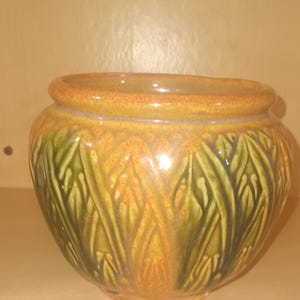 May include: A ceramic bowl with a wide rim and a rounded body. The bowl is decorated with a leaf-like pattern in shades of green, orange, and brown. The rim is a darker shade of brown.