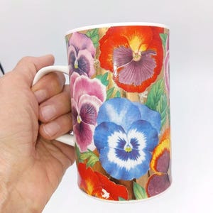 May include: A white ceramic mug featuring a vibrant floral pattern of pansies in shades of red, blue, purple, and pink, set against a metallic gold background. The mug has a simple white handle and a cylindrical shape with a smooth, glossy finish.