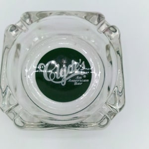 May include: A square, clear glass ashtray featuring a circular dark green emblem in the center. The emblem displays the white text Clyde's and the smaller text An American Bar. The ashtray has four indented notches along its thick, molded glass rim.