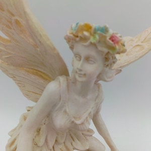 May include: A cream-colored fairy figurine featuring large, detailed wings and a floral crown with pastel pink, blue, and yellow accents. The figure is depicted in a kneeling pose, wearing a layered dress with a textured, ruffled skirt design.