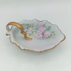 May include: A white porcelain shell-shaped dish featuring a hand-painted cluster of pink roses and green leaves. The dish has a gold-painted handle and a gold-trimmed scalloped edge. The signature Louise Faulkner is visible in black script.