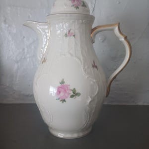 May include: A white porcelain coffee pot with a lid, handle, and spout. The pot is decorated with pink roses and gold accents. The lid is topped with a gold finial. The pot has a classic, elegant design.
