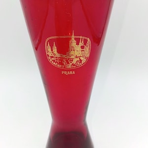 May include: A tall, tapered ruby red glass vase featuring a gold-colored graphic illustration of the Hradčany castle complex in Prague. The text "HRADČANY" is printed in a curved arc within the oval graphic, with the word "PRAHA" below it.