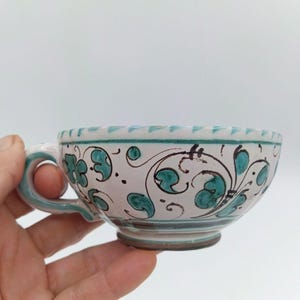 May include: A ceramic teacup featuring a white base with hand-painted teal floral and vine motifs. The cup has a curved handle, a wide rim with a teal triangular pattern, and a dark brown base. The exterior is decorated with swirling lines.