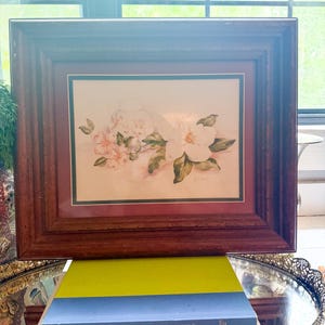 May include: A rectangular wooden picture frame containing a botanical print of white flowers and green leaves, set against a dark red mat. The frame rests on two stacked books, one with a blue spine titled The SHABBY CHIC Home and one white.