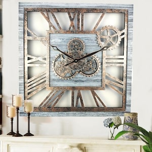 Puede incluir: Un reloj de pared cuadrado grande con un marco de madera blanca desgastada y marcas de números romanos. La esfera del reloj presenta un diseño de engranajes y una manecilla de reloj negra.