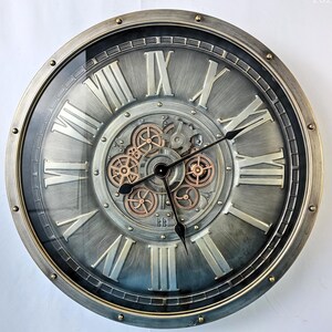 May include: A large, round wall clock with a metallic gray finish and Roman numerals. The clock features visible gears in the center and black clock hands. The outer rim has a gold-colored trim. The clock's design has a vintage, industrial aesthetic.