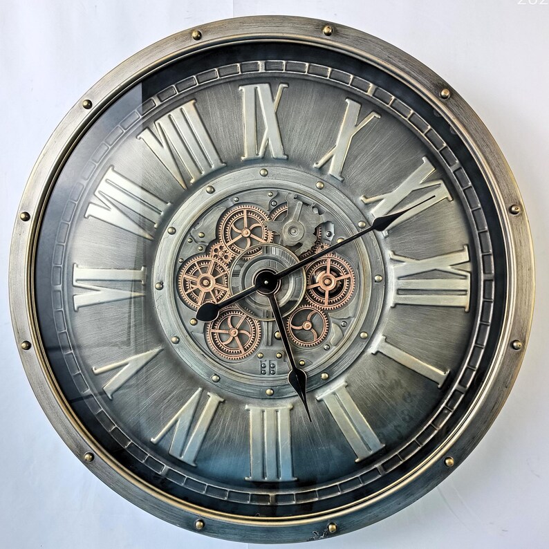 May include: A large, round wall clock with a metallic gray finish and Roman numerals. The clock features visible gears in the center and black clock hands. The outer rim has a gold-colored trim. The clock's design has a vintage, industrial aesthetic.