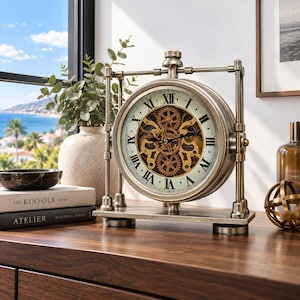 May include: A round table clock with a metallic silver frame and a skeleton dial featuring exposed gold-toned gears. The clock face displays black Roman numerals and two black hands, supported by a rectangular base with vertical side pillars.