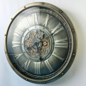 May include: A large, round wall clock with a metallic gray face and a bronze-colored frame. The clock features Roman numerals and visible gears in the center. The hands are black, and the clock has a glass cover.