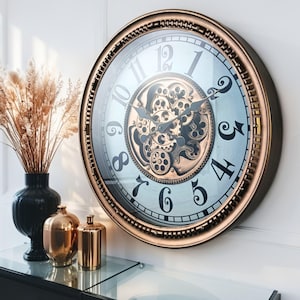 May include: A large, round wall clock with a bronze-colored frame and a light blue face. The clock features black numerals and intricate gear details in the center. The clock is mounted on a white wall.