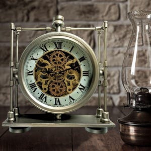 May include: A silver and gold clock with exposed gears on a metal stand. The clock face has Roman numerals and a white background.