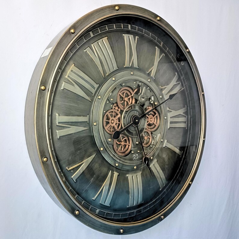 May include: A large, round wall clock with a metallic gray finish and gold accents. The clock face features Roman numerals and visible gears in the center. The clock hands are black.