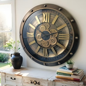 May include: Large, round wall clock with a bronze-colored metal frame and Roman numerals. The clock face features visible gears and a glass cover. The clock is mounted on a white wall above a white wooden cabinet. A black vase with greenery is on the cabinet.