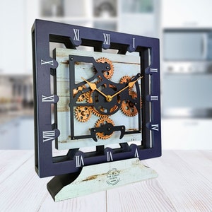 May include: A black and white clock with a blue and white wooden base. The clock face features a gear design with gold accents. The clock is labeled "The Gears Clock".