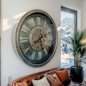 May include: Large, round wall clock with a metallic gray frame and a distressed teal face. The clock features Roman numerals and visible gears in the center. The clock is mounted on a white wall above a brown leather sofa.