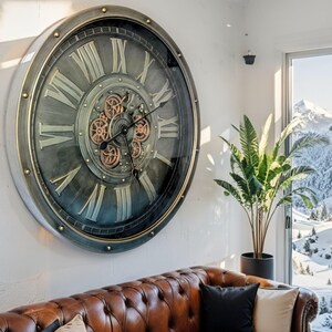 May include: Large, round wall clock with a metallic gray and gold finish. The clock features Roman numerals and visible gears in the center. The clock is mounted on a white wall above a brown leather sofa. The clock's diameter is approximately 40 inches.
