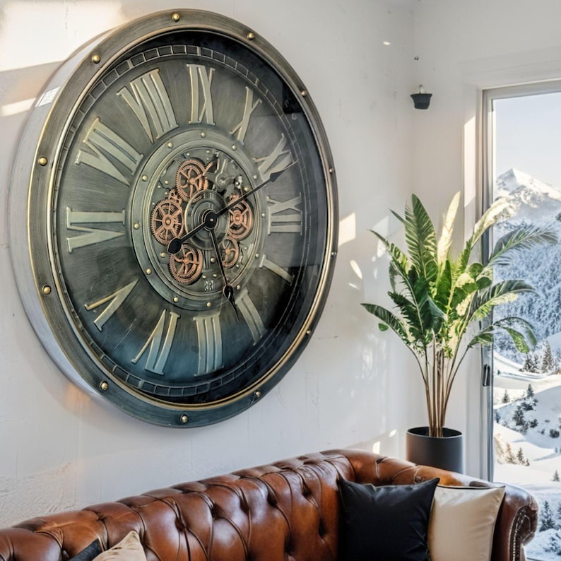 May include: Large, round wall clock with a metallic gray and gold finish. The clock features Roman numerals and visible gears in the center. The clock is mounted on a white wall above a brown leather sofa. The clock's diameter is approximately 40 inches.
