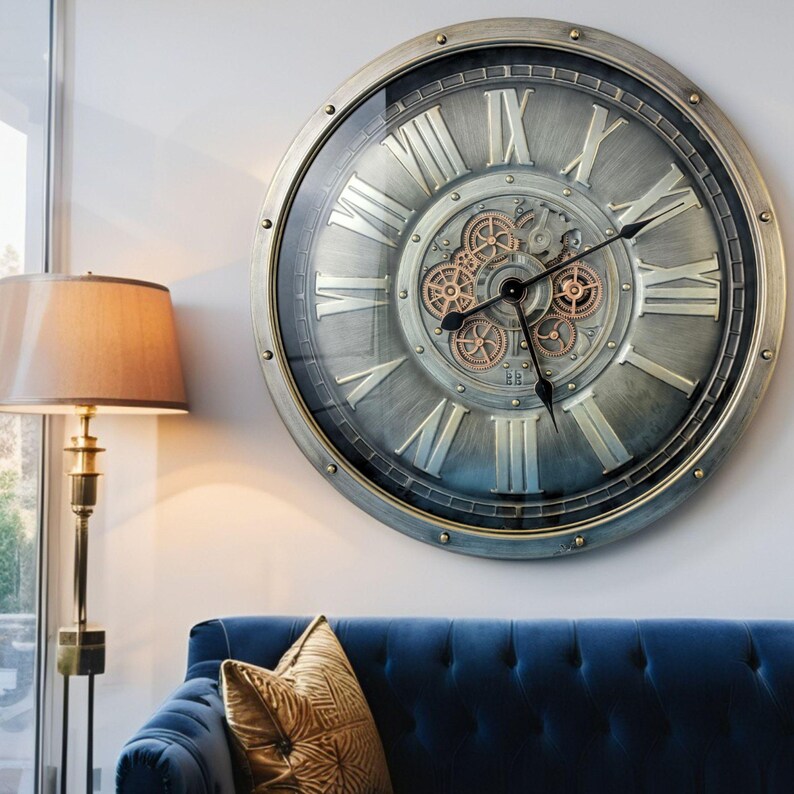 May include: Large, round wall clock with a metallic gray finish and Roman numerals. The clock features visible gears in the center and a glass cover. The clock's frame is a darker gray with rivets. The clock is mounted on a white wall.