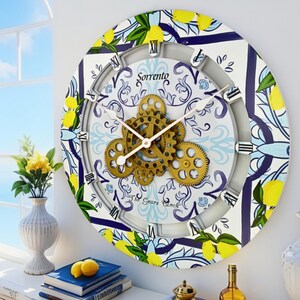 May include: A round wall clock featuring a white background with blue floral patterns and yellow lemon illustrations. The center displays exposed gold-colored mechanical gears, Roman numerals, and the text Sorrento and The Gears Clock.