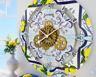May include: A round wall clock featuring a white background with blue floral patterns and yellow lemon illustrations. The center displays exposed gold-colored mechanical gears, Roman numerals, and the text Sorrento and The Gears Clock.