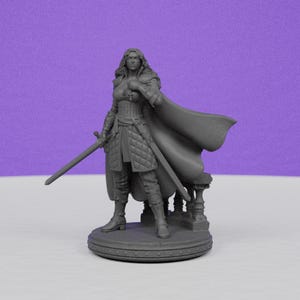 May include: A gray figurine of a female warrior, holding a sword and wearing a cape. The figure is standing on a round base with a decorative edge. The warrior is dressed in armor and boots, with long flowing hair.