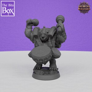 May include: A gray miniature figurine of a fantasy creature with a drum on its chest and two drumsticks raised in the air. The figurine has a menacing expression and is standing on a small base. The Bits Box and Daybreak Miniatures logos are visible.